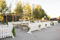 Tuska Festivaalin rakennusvaiheessa kesällä 2024 otettu kuva valkosiesta puuaidasta ja kevyistä viherkasvisomisteista Valkoista puuaitaa ja penkkipöytäsettejä kesäisessä illassa Helsingin Suvilahdessa (Tuska Festivaalin rakennusvaihe)